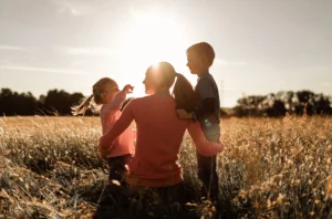 Mom With Two Young Children, Playing In A Field Of Tall Grass At Sunset.