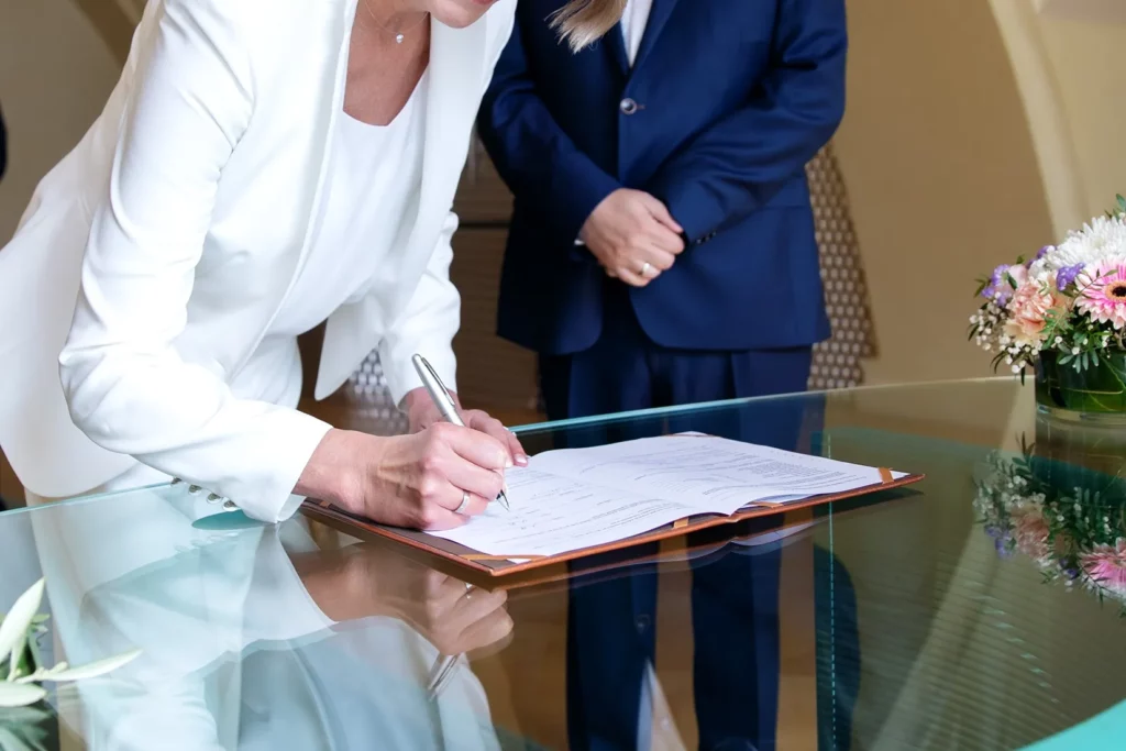 A Person Wearing A White Pantsuit Signing An Agreement With A Lawyer Standing In The Background.