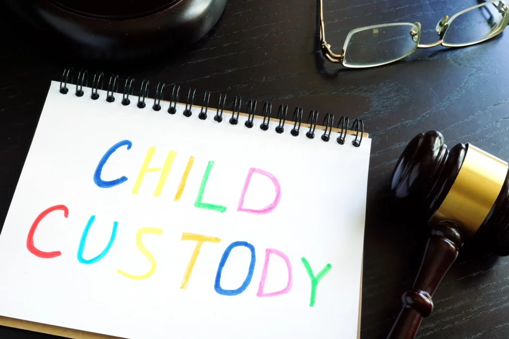 A Notepad With The Word 'Child Custody' Written In Multi Colored Markers, A Gavel And A Pair Of Eyeglasses Sitting On A Desk.